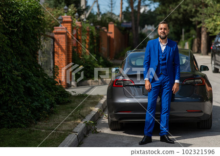 Caucasian bearded man in a blue suit stands near a black car in the countryside in summer. 102321596