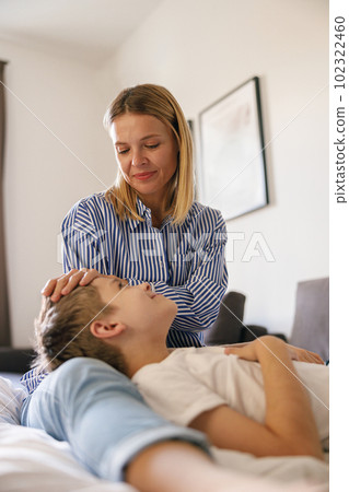 Young boy lying on mother's lap on sofa in living room at home while woman touches the child's hair 102322460