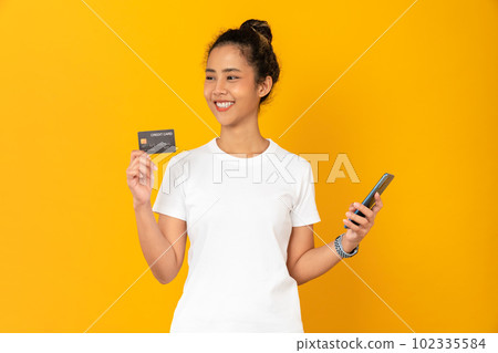 Cheerful young Asian woman holding smartphone with show mockup credit card on yellow background. 102335584