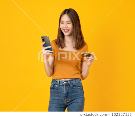Cheerful young Asian woman holding smartphone with show mockup credit card on yellow background. Cheerful young Asian woman holding smartphone with show mockup credit card on yellow background. 102337135