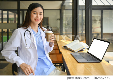 Portrait of asian female doctor in white uniform and stethoscope holding paper cup of coffee sitting hospital canteen 102337359