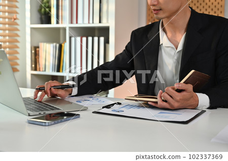 Male economist in black suit preparing report and analyzing work results at workstation Male economist in black suit preparing report and analyzing work results at workstation 102337369