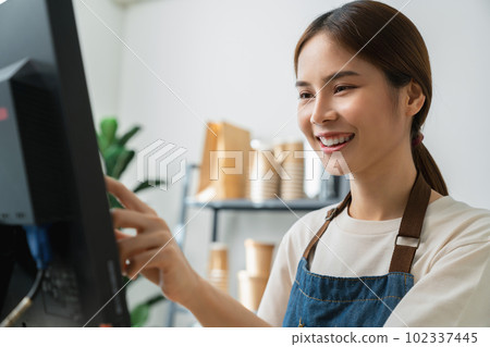 Asian woman cashier wears an apron and using pos terminal to input orders on coffee shop counter. 102337445