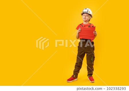 Full-length portrait of boy imitating worker. Child wears helmet with holds toolbox, isolation on yellow background. Copy space, mock up Full-length portrait of boy imitating worker. Child wears helmet with holds toolbox, isolation on yellow background. Copy space, mock up 102338293