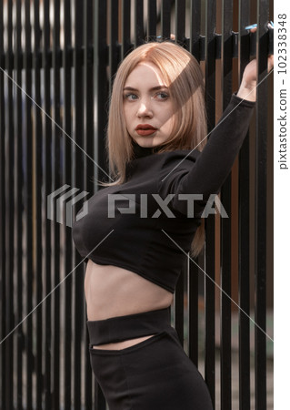 Stylish young beauty with beautiful figure in short black top and bare stomach. Blonde girl poses near metal fence. Vertical frame. 102338348