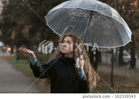 Portrait of young woman standing under transparent umbrella to see if it is raining. Girl with brown hair walks in park Portrait of young woman standing under transparent umbrella to see if it is raining. Girl with brown hair walks in park 102338543