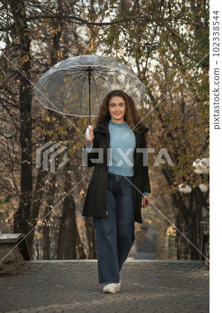 Pretty young woman with lush hair standing with transparent umbrella in autumn park. Photo of woman in park Pretty young woman with lush hair standing with transparent umbrella in autumn park. Photo of woman in park 102338544