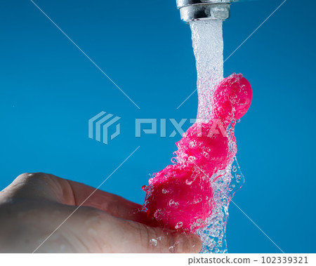 Woman holding pink anal beads under running water on blue background. Sex toy hygiene concept. Woman holding pink anal beads under running water on blue background. Sex toy hygiene concept. 102339321