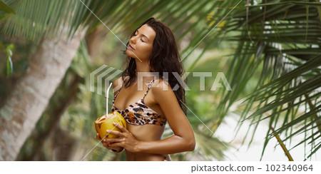 Beach Babe, Tanned Bikini-Clad Woman Enjoying Fresh Coconut Water on Sandy Shore 102340964