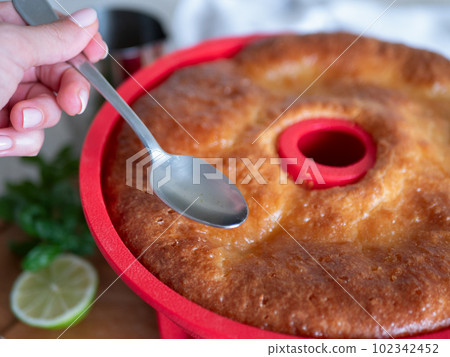 Freshly-baked Savarin in red silicone cake mold is poured by syrup. Rum-soaked circular cake, close up Freshly-baked Savarin in red silicone cake mold is poured by syrup. Rum-soaked circular cake, close up 102342452