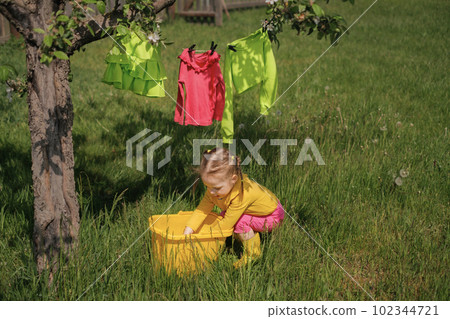 The child takes out wet clothes after laundry from a bucket and hangs them on a clothesline to dry in the wind. Washing powder for hand washing of bright multicolored underwear The child takes out wet clothes after laundry from a bucket and hangs them on a clothesline to dry in the wind. Washing powder for hand washing of bright multicolored underwear 102344721