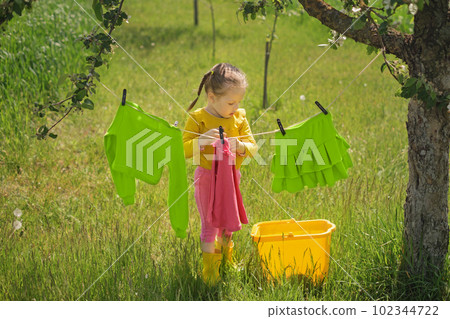 A girl with pigtails hangs a T-shirt on a clothesline with a clothespin. Washing powder and conditioner for hand and machine laundry of bright colored fabrics. A girl with pigtails hangs a T-shirt on a clothesline with a clothespin. Washing powder and conditioner for hand and machine laundry of bright colored fabrics. 102344722