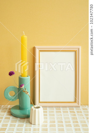 Empty picture frame and candle, flower on check tile table. yellow wall background 102347780