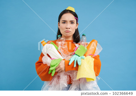 Angry young woman, holding empty plastic bottles and garbage, sorting waste to recycle, looks upset by unsorted trash in her hands, blue background Angry young woman, holding empty plastic bottles and garbage, sorting waste to recycle, looks upset by unsorted trash in her hands, blue background 102366649