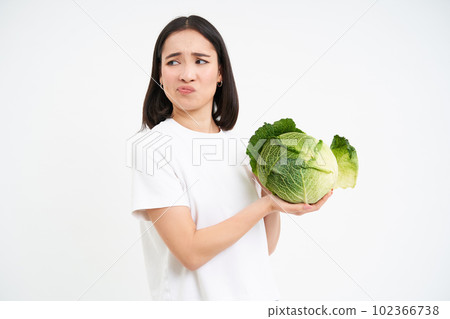 Asian woman looks with disgust and dislike at cabbage, hates vegetables, isolated on white background Asian woman looks with disgust and dislike at cabbage, hates vegetables, isolated on white background 102366738