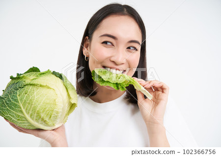 Close up of cute korean woman, eating cabbage, on diet, holding lettuce, isolated on white background Close up of cute korean woman, eating cabbage, on diet, holding lettuce, isolated on white background 102366756