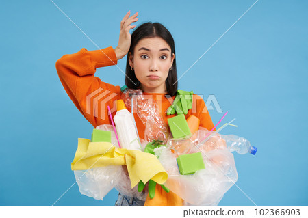 Confused Korean woman tries to go green, recycle plastic household items, holding bottles and gloves, does recycling, blue background Confused Korean woman tries to go green, recycle plastic household items, holding bottles and gloves, does recycling, blue background 102366903