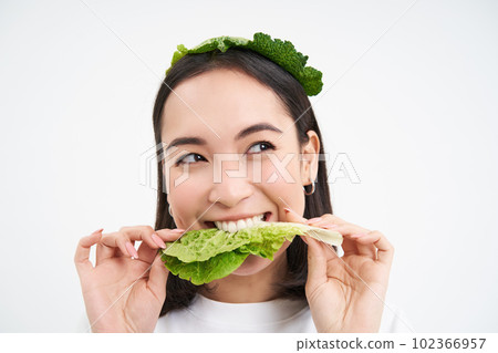 Close up portrait of smiling asian woman eating lettuce, loves cabbage, vegetarian enjoys raw organic food, white background 102366957