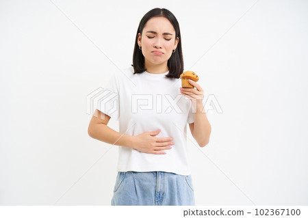Portrait of korean woman has stomach ache, touching belly, holding cupcake, cant eat pastry, white studio background Portrait of korean woman has stomach ache, touching belly, holding cupcake, cant eat pastry, white studio background 102367100