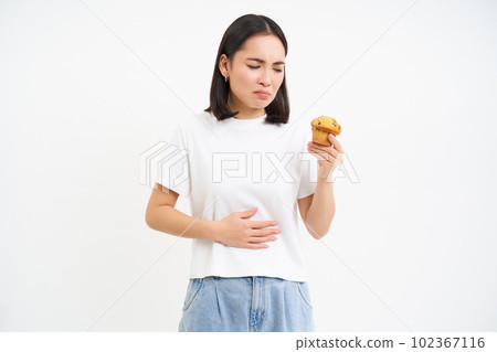 Portrait of asian woman with stomach ache, holding cupcake, food poisoning, feeling unwell, studio background Portrait of asian woman with stomach ache, holding cupcake, food poisoning, feeling unwell, studio background 102367116