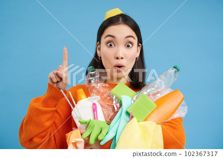 Happy young woman pointing up, holding plastic empty bottles and rubbish for recycling, showing eco banner, sorting station advertisement, blue background 102367317