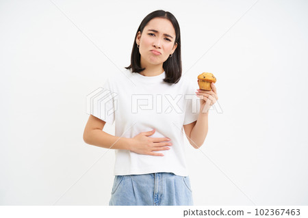 Portrait of asian woman with stomach ache, holding cupcake, food poisoning, feeling unwell, studio background Portrait of asian woman with stomach ache, holding cupcake, food poisoning, feeling unwell, studio background 102367463