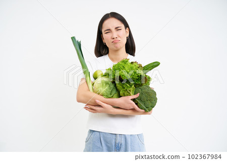 Woman with disgusted face, holding vegetables, green organic food, hates diets and detox, white background 102367984
