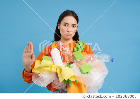 Serious eco activist shows stop sign and plastic waste, urges to stop throwing recycling materials into general garbage bins, blue background Serious eco activist shows stop sign and plastic waste, urges to stop throwing recycling materials into general garbage bins, blue background 102368005