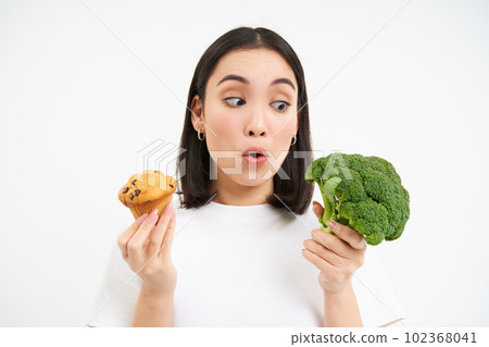 Portrait of asian woman looks excited at broccoli, holding cupcake, white background Portrait of asian woman looks excited at broccoli, holding cupcake, white background 102368041