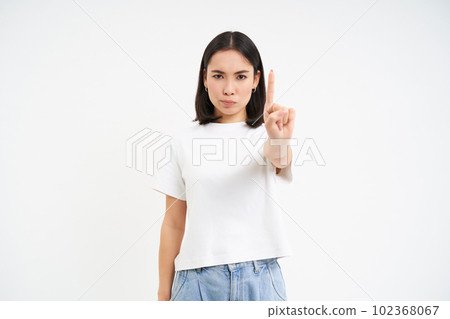 Serious 25 years woman, shows one finger, stop sign, warning gesture, disapprove smth, stands over white background Serious 25 years woman, shows one finger, stop sign, warning gesture, disapprove smth, stands over white background 102368067