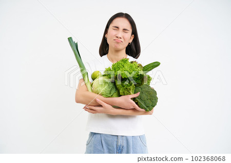Miserable asian woman, holds green vegetables and complains at het diet, looks at calories, white background 102368068