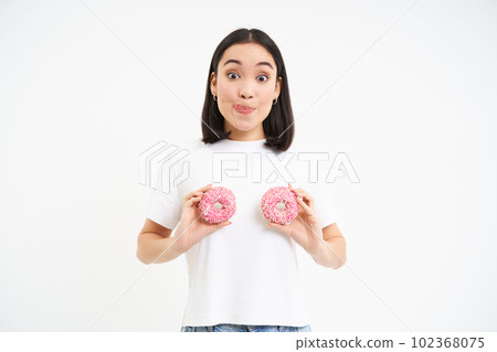 Smiling happy asian woman laughing, holding two glazed doughnuts over chest, standing over white background 102368075