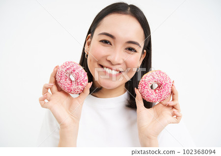 Close up portrait of cute korean woman, smiling while eating pink glazed doughnuts, likes tasty donnuts, white background 102368094