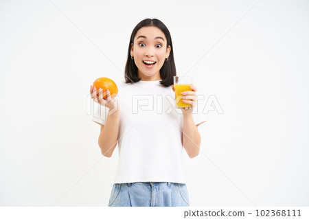 Image of korean woman, looks surprised at camera, holds fresh juice and orange fruit in hands, white background 102368111