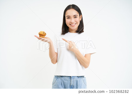 Portrait of smiling young woman pointing finger at cupcake, showing her favourite bakery dessert, white background Portrait of smiling young woman pointing finger at cupcake, showing her favourite bakery dessert, white background 102368282