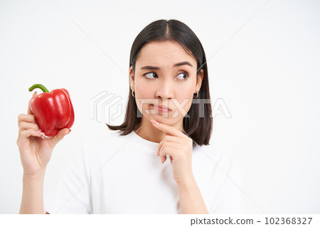 Close up portrait of asian woman, holding red sweet pepper and thinking, stands with pensive face, white background 102368327