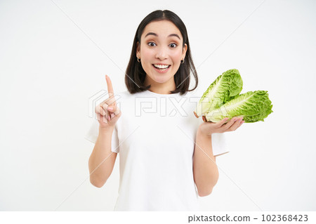 Image of smiling asian girl shows cabbage and one finger, points, teaches healthy diet, stands over white background with lettuce Image of smiling asian girl shows cabbage and one finger, points, teaches healthy diet, stands over white background with lettuce 102368423