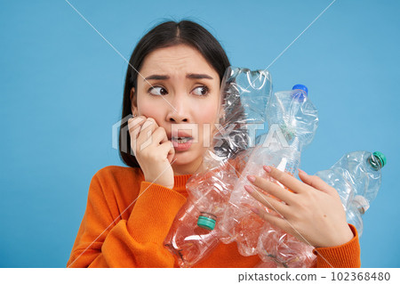 Portrait of asian girl with scared face, holding piles of plastic bottles for recycling and stares to the right with shocked emotion, blue backgrond 102368480