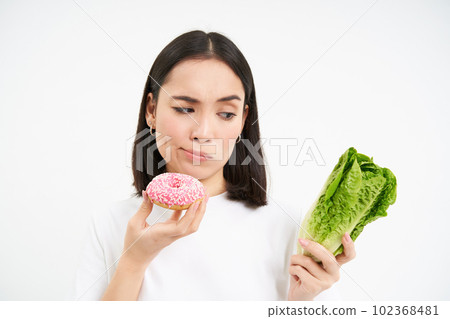 Healthy clean detox eating concept. Vegetarian, vegan, raw concept. Woman eats tasty doughnut and looks skeptical at cabbage vegetable, white background 102368481