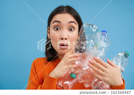 Surprised korean woman, holding plastic bottles, looking shocked at camera, concept of environment and recycling, blue background 102368487