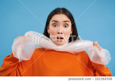 Portrait of young woman being choked by plastic bags, concept of recycling and environment, blue background 102368506