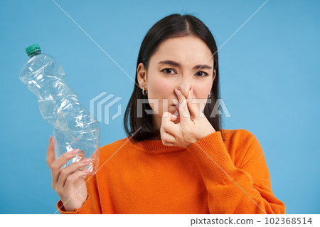Portrait of korean girl shuts nose, shows plastic bottle, stinky trash, stands over blue background Portrait of korean girl shuts nose, shows plastic bottle, stinky trash, stands over blue background 102368514