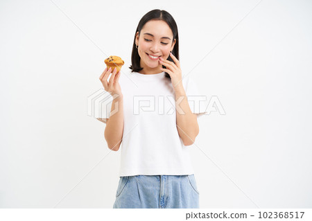Bakery and sweets. Happy asian woman looking at tasty cupcake, eating pastry, white background Bakery and sweets. Happy asian woman looking at tasty cupcake, eating pastry, white background 102368517