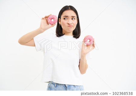 Image of asian girl holds two doughnuts, thinking of breaking her diet, looks with doubt at donnuts, white background Image of asian girl holds two doughnuts, thinking of breaking her diet, looks with doubt at donnuts, white background 102368538