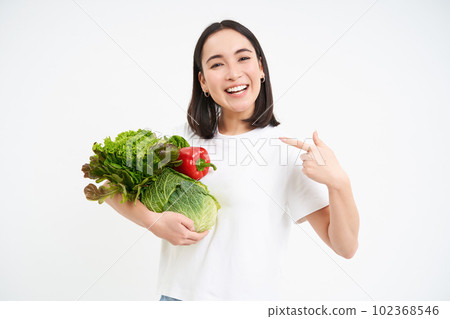 Image of happy korean woman, pointing at vegetables, smiling, recommending eating healthy raw food, green diet concept, white background Image of happy korean woman, pointing at vegetables, smiling, recommending eating healthy raw food, green diet concept, white background 102368546