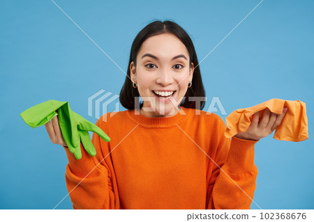 Enthusiastic asian woman ready to clean her apartment, showing two different latex gloves and smiling, blue background 102368676