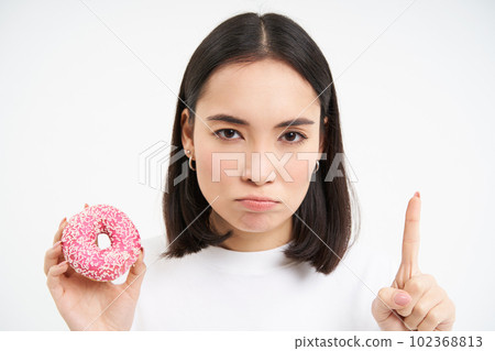 Close up of serious asian woman, shows taboo, stop gesture and pink glazed doughnut, forbids eating sweets, white background Close up of serious asian woman, shows taboo, stop gesture and pink glazed doughnut, forbids eating sweets, white background 102368813