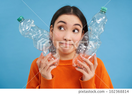 Thoughtful korean woman shows two plastic bottles, looks clueless and puzzled, concept of recycling and ecology Thoughtful korean woman shows two plastic bottles, looks clueless and puzzled, concept of recycling and ecology 102368814
