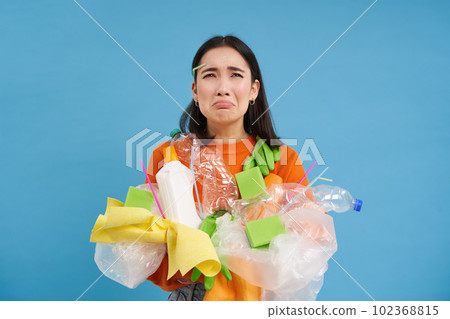 Crying asian female holding plastic waste, garbage in hands and looking upset, has to recycle, blue background Crying asian female holding plastic waste, garbage in hands and looking upset, has to recycle, blue background 102368815