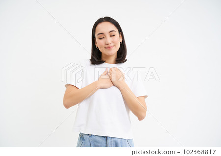 Smiling young korean woman, holds hands on heart, concept of love, sympathy, kindness, isolated on white background Smiling young korean woman, holds hands on heart, concept of love, sympathy, kindness, isolated on white background 102368817
